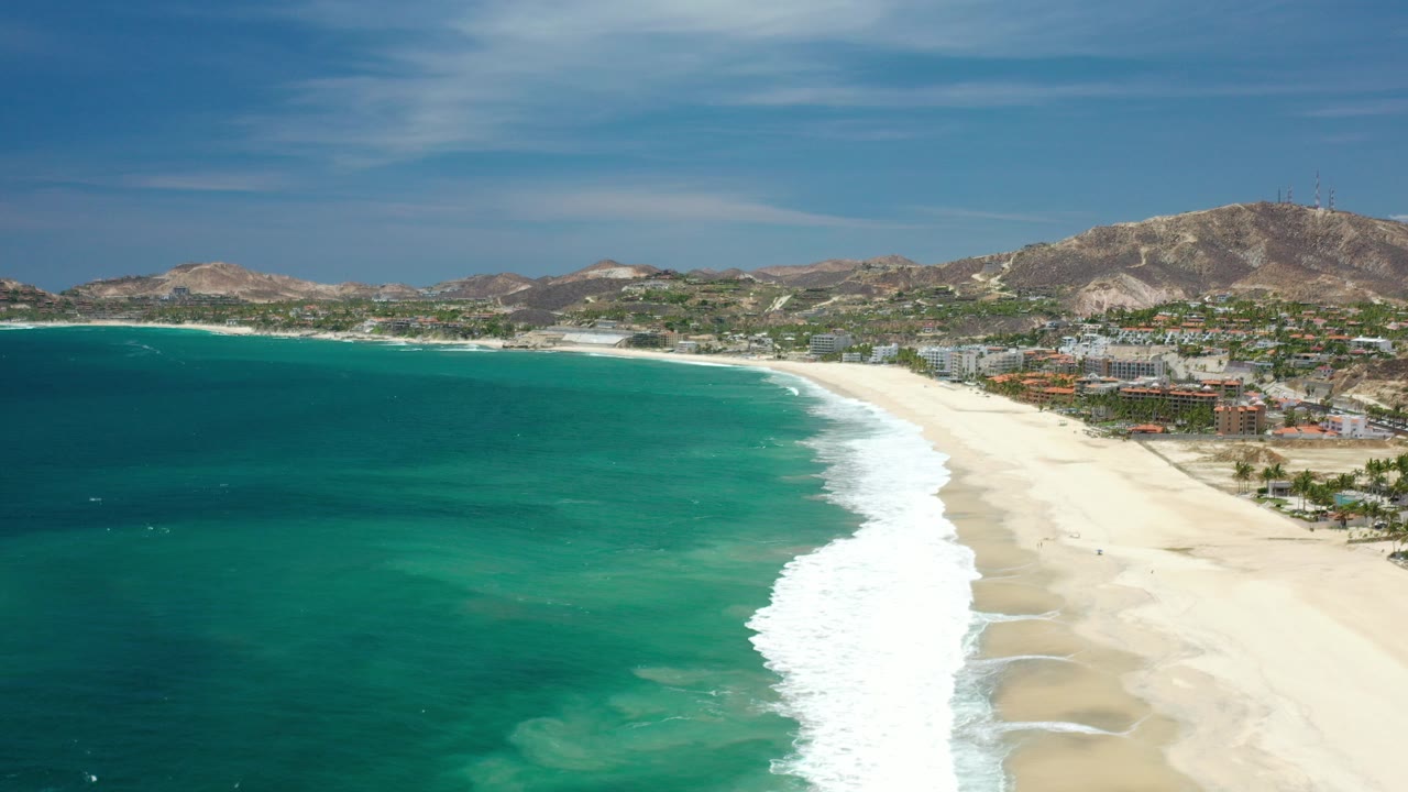 Aerial view in Los Cabos, Mexico with turquoise ocean on the left, a bright surf line on the beach, distant coastal buildings, dry hills, and no visible people.