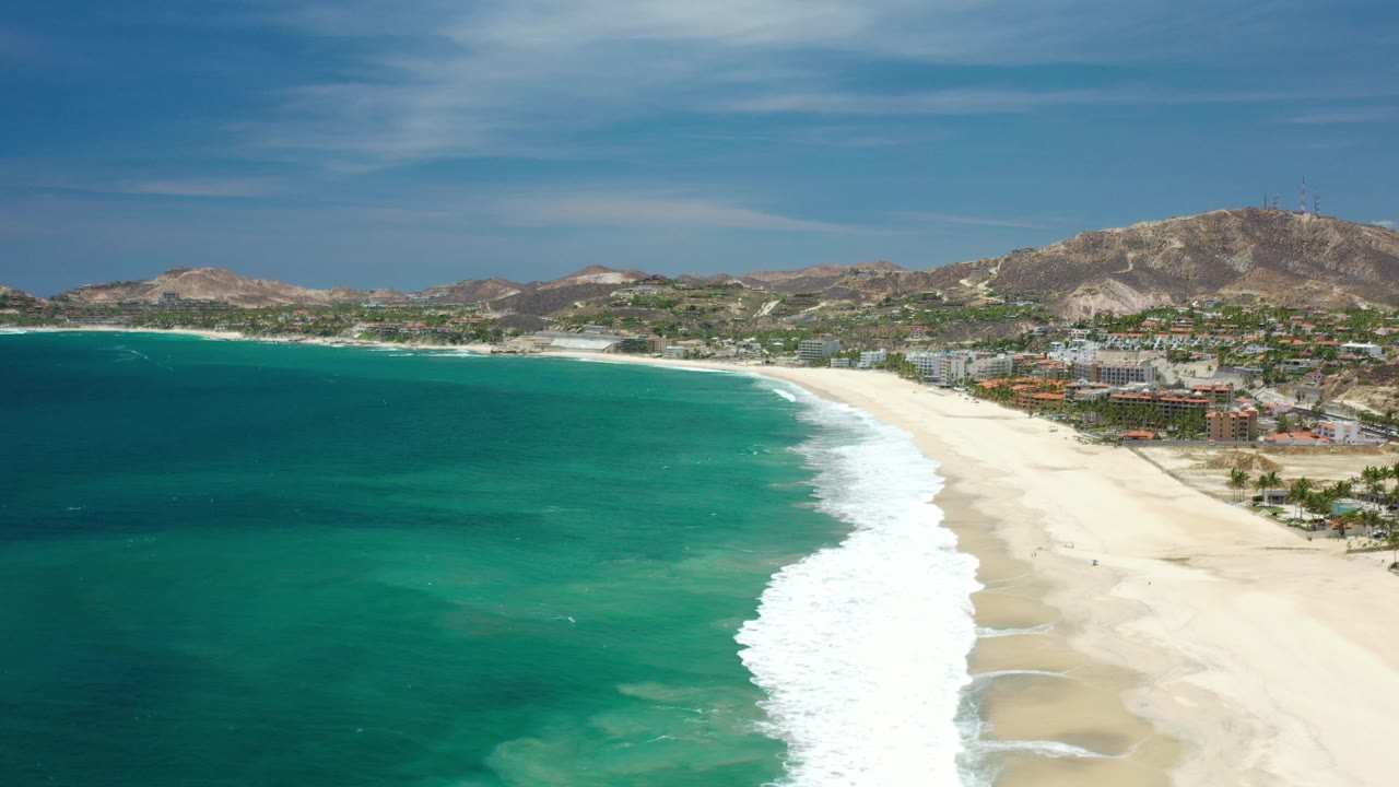 Drone-style view of turquoise sea, curling surf, long beach, buildings, and hills under blue sky.