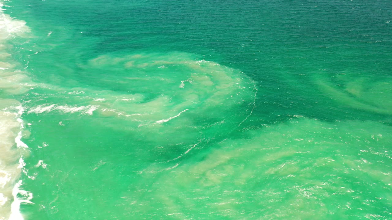 Aquamarine water off Los Cabos, Mexico, shows a pale suspended sand swirl near center with white surf foam along the left edge and no visible people.