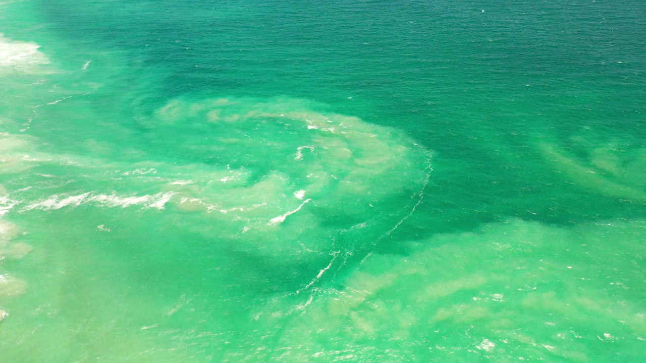 Turquoise water with pale sand clouds and a curved foam line.