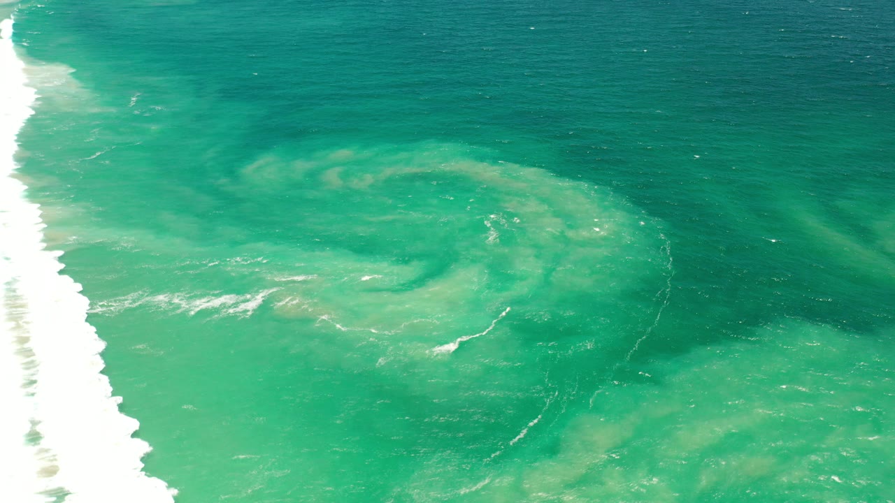 Overhead view of teal ocean water in Los Cabos, Mexico, with a bright white surf band at left, pale underwater swirls near center, and no visible people.