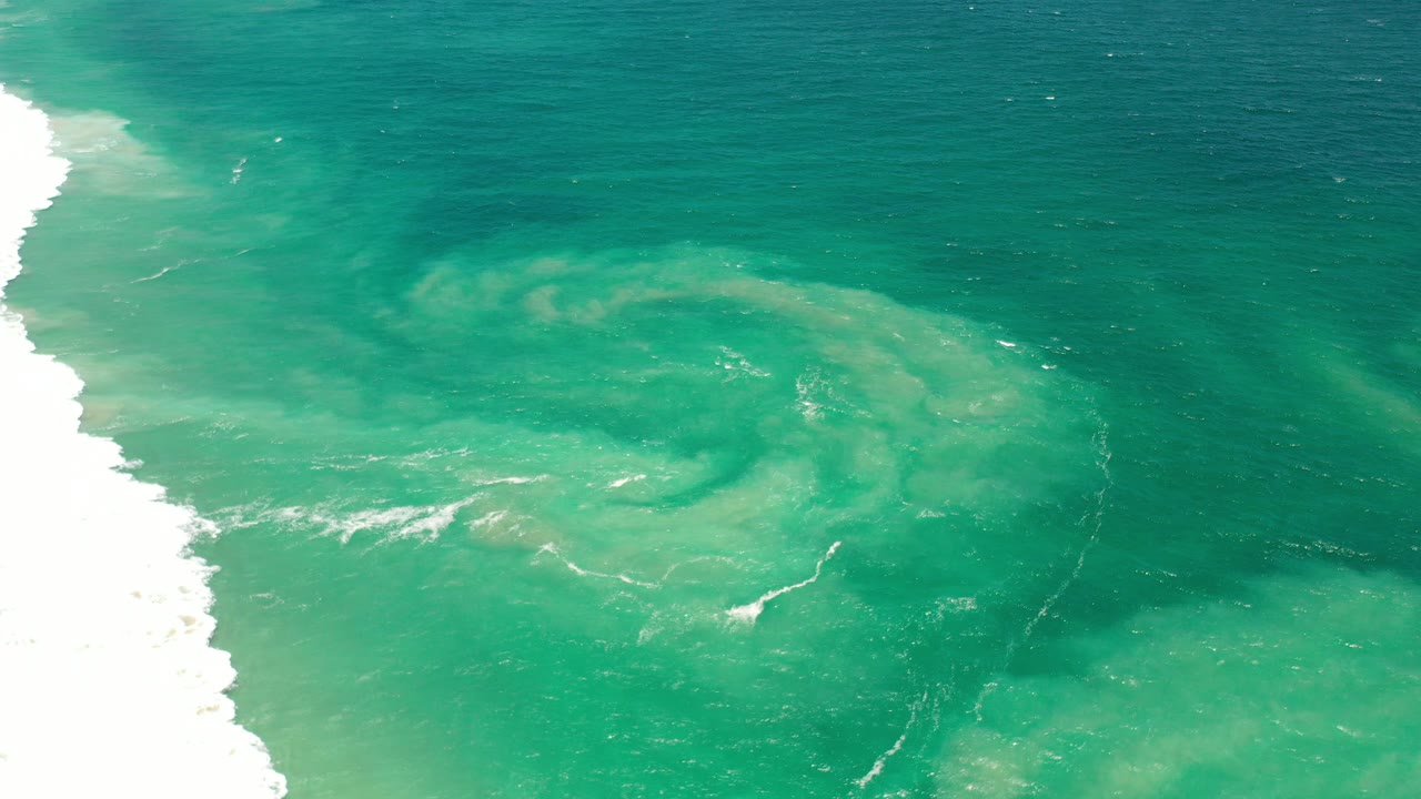 Elevated view of green sea off Los Cabos, Mexico, with white breaking surf at the left edge and a pale foam swirl in the center, no visible people.
