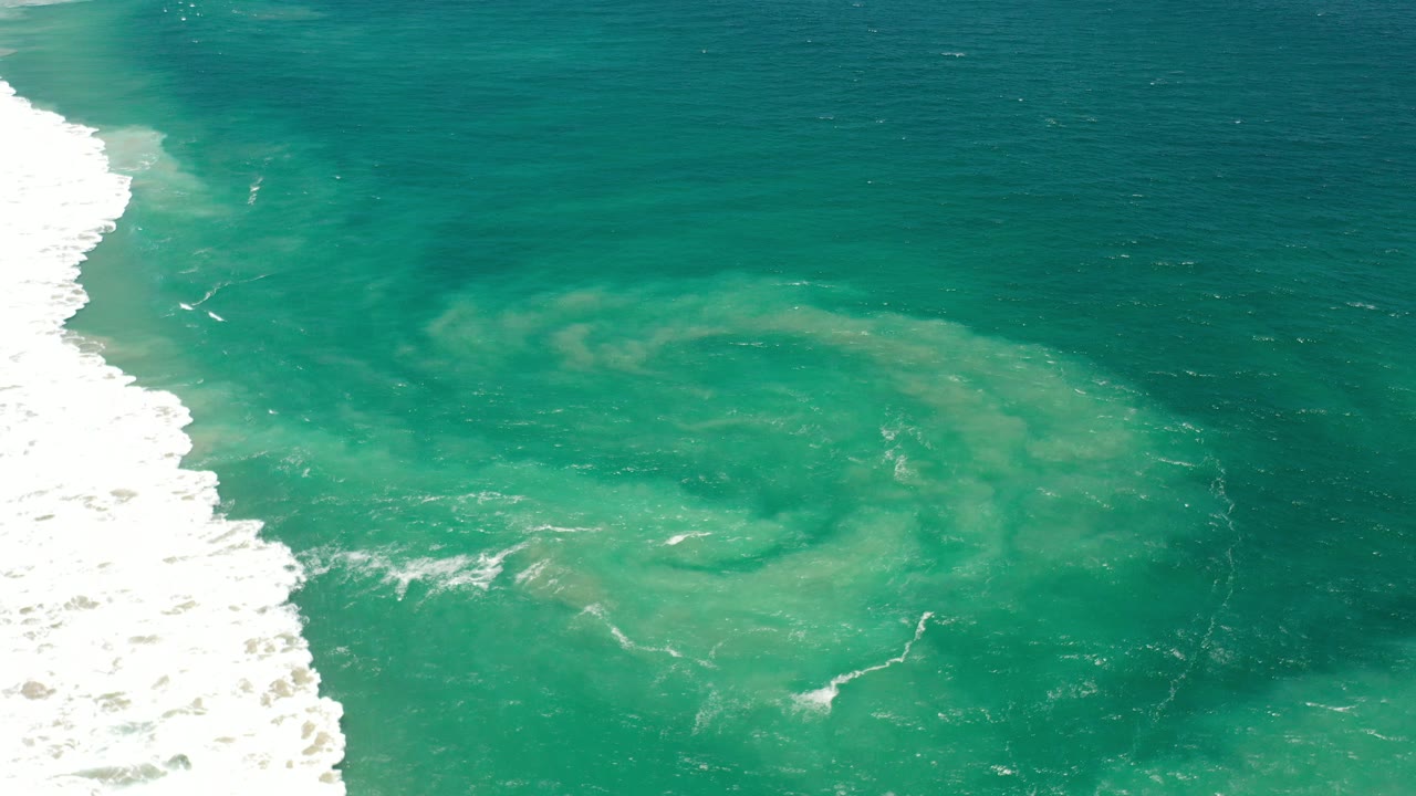 Aerial-looking view of green ocean water in Los Cabos, Mexico with a pale circular swirl near the center, white surf along the left edge, and no visible people.