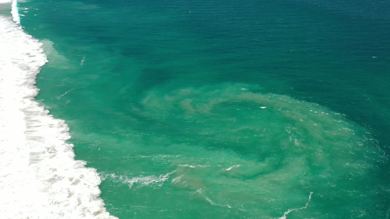 Aerial-style frame of Los Cabos ocean water with a bright foam band on the left and a pale submerged patch spreading through the center, no visible people.