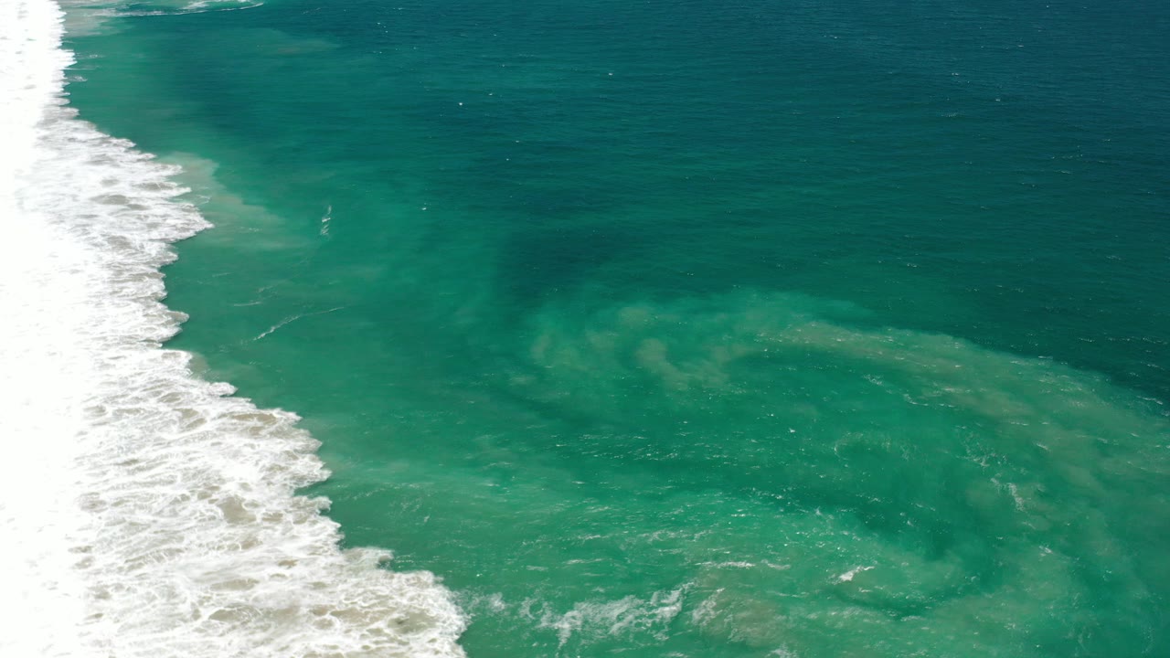 White surf enters from the left edge beside teal Los Cabos ocean water, with darker current shapes under the surface and no visible people.