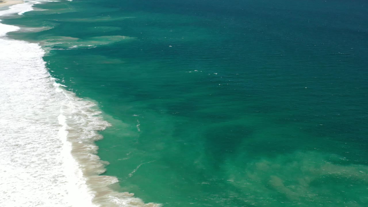 Los Cabos aerial shoreline frame with a broad band of white sea foam at left, green-blue shallow water near center, darker ocean at right, and no visible people.