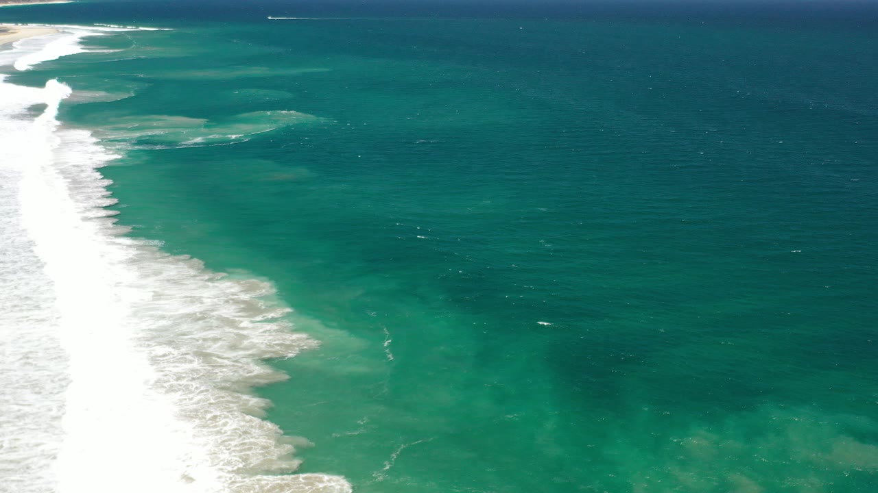 Aerial frame from Los Cabos, Mexico showing white surf along the left side, blue green water across the frame, a thin horizon, and no visible people.