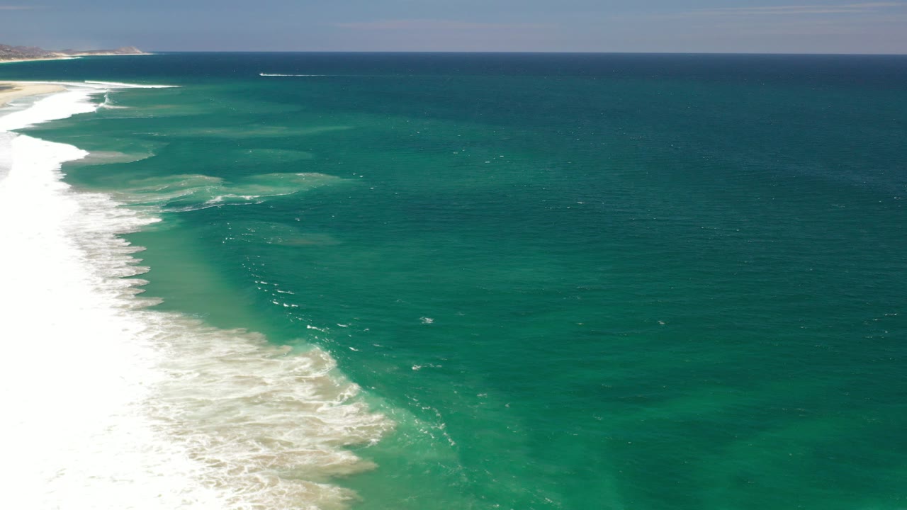 Aerial side view of the Los Cabos, Mexico coastline with white surf along the left edge, broad blue-green ocean to the right, a level horizon, and no visible people.