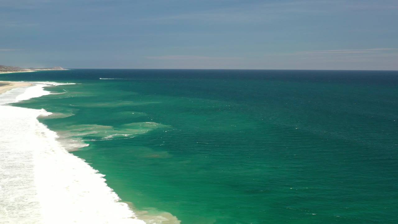 Opening frame of Los Cabos, Mexico coastline with white surf along the left edge, green shallow water, deeper blue ocean, distant land on the horizon, and no visible people.