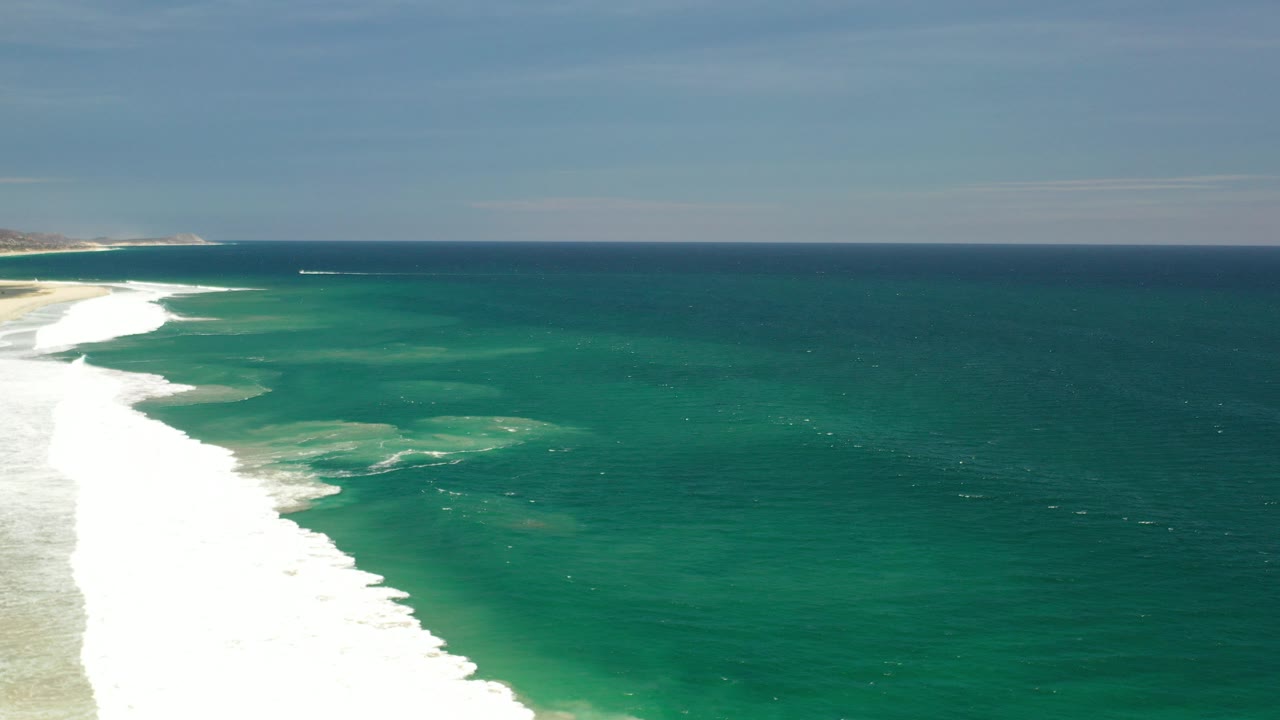 Opening aerial frame over the Los Cabos shoreline with white surf at the left edge, teal ocean to the right, a distant coast on the horizon, and no visible people.