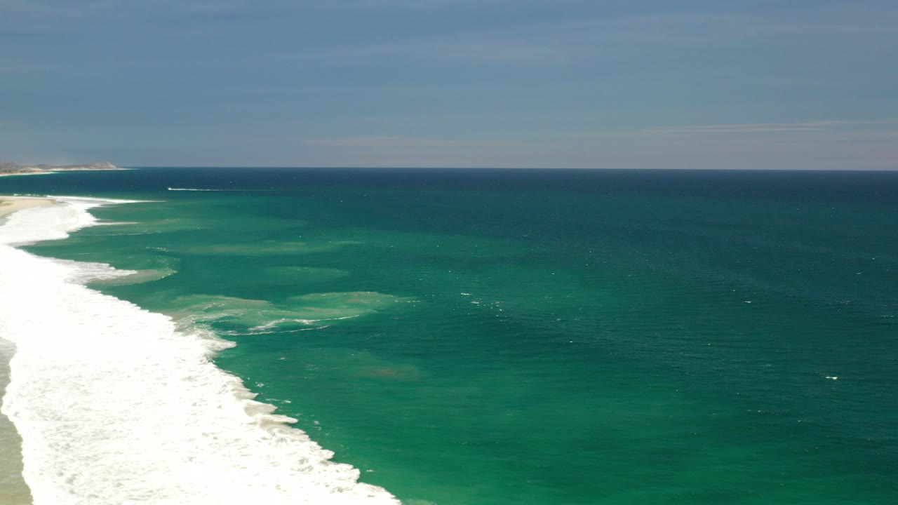 Opening aerial frame of a Los Cabos shoreline with white surf on the left, green ocean water across the center, a distant horizon, and no visible people.