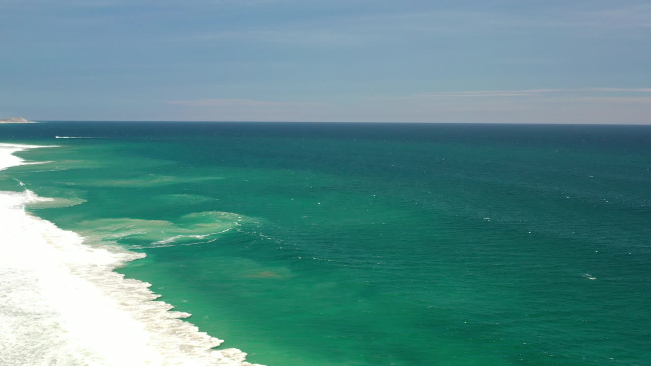 Opening frame of a Los Cabos, Mexico aerial ocean view, with white surf entering from the lower left, turquoise water across the frame, a flat horizon, and no visible people.
