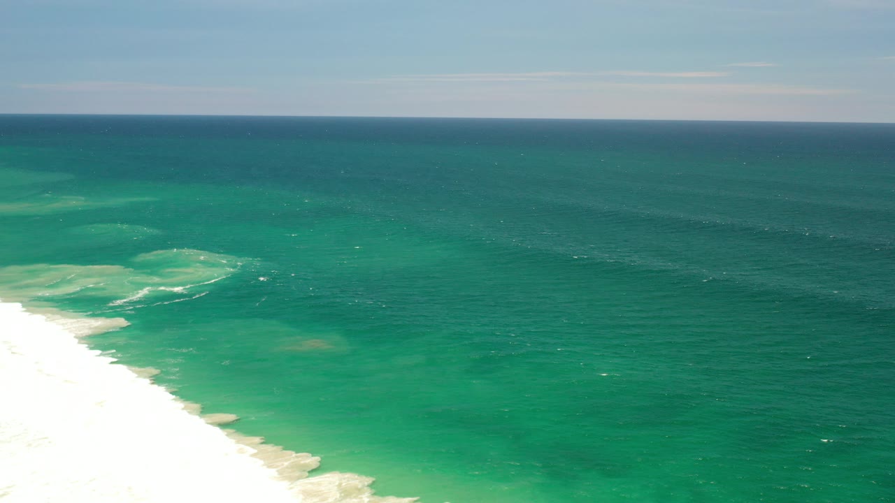 Aerial view over Los Cabos, Mexico, with green-blue ocean, white surf along the left beach edge, a level horizon, pale sky, and no visible people.