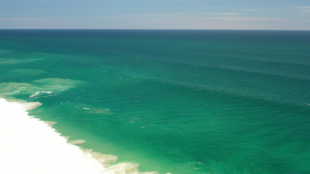 Opening frame of the Los Cabos, Mexico coastline with blue green ocean, white surf entering from the left edge, a pale overcast sky, and no visible people.