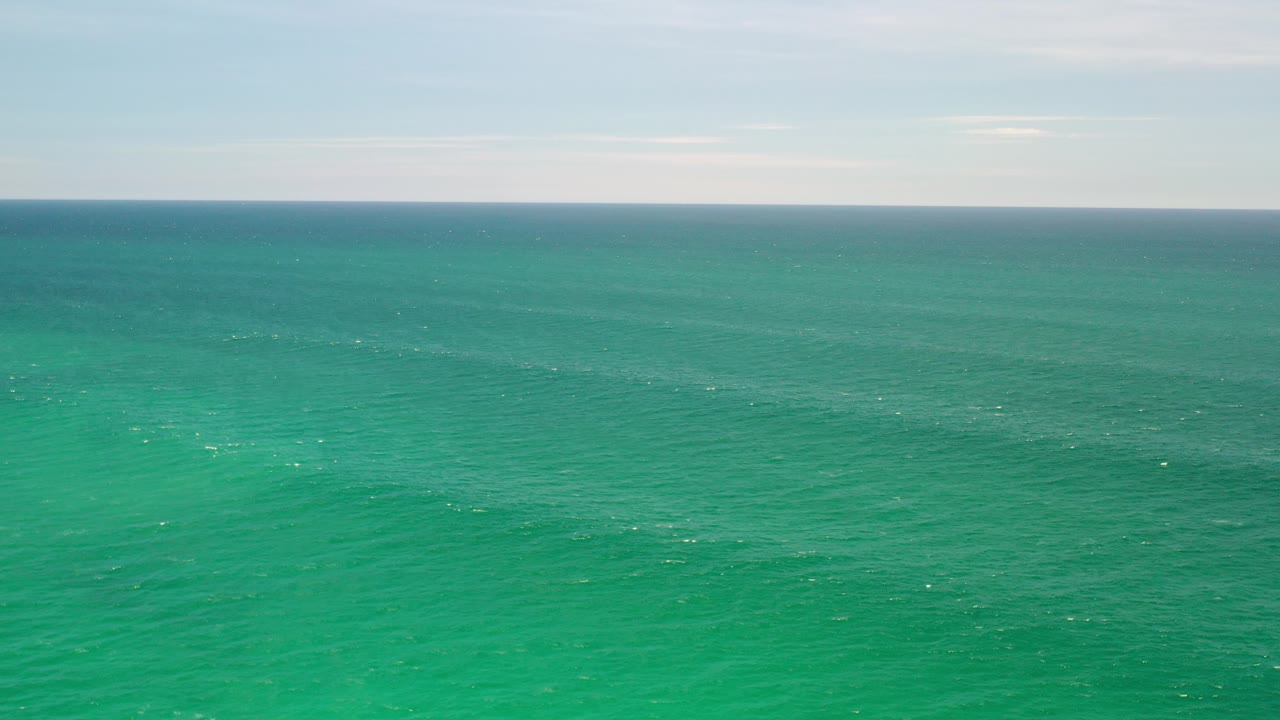 Wide aerial view of green ocean water in Los Cabos, Mexico, with a pale sky, distant level horizon, scattered white wave flecks, and no visible people.