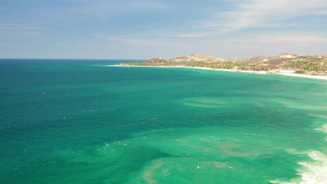 Aerial view over Los Cabos, Mexico, with turquoise ocean in the foreground, a sandy shoreline and low hills in the distance, and no visible people.