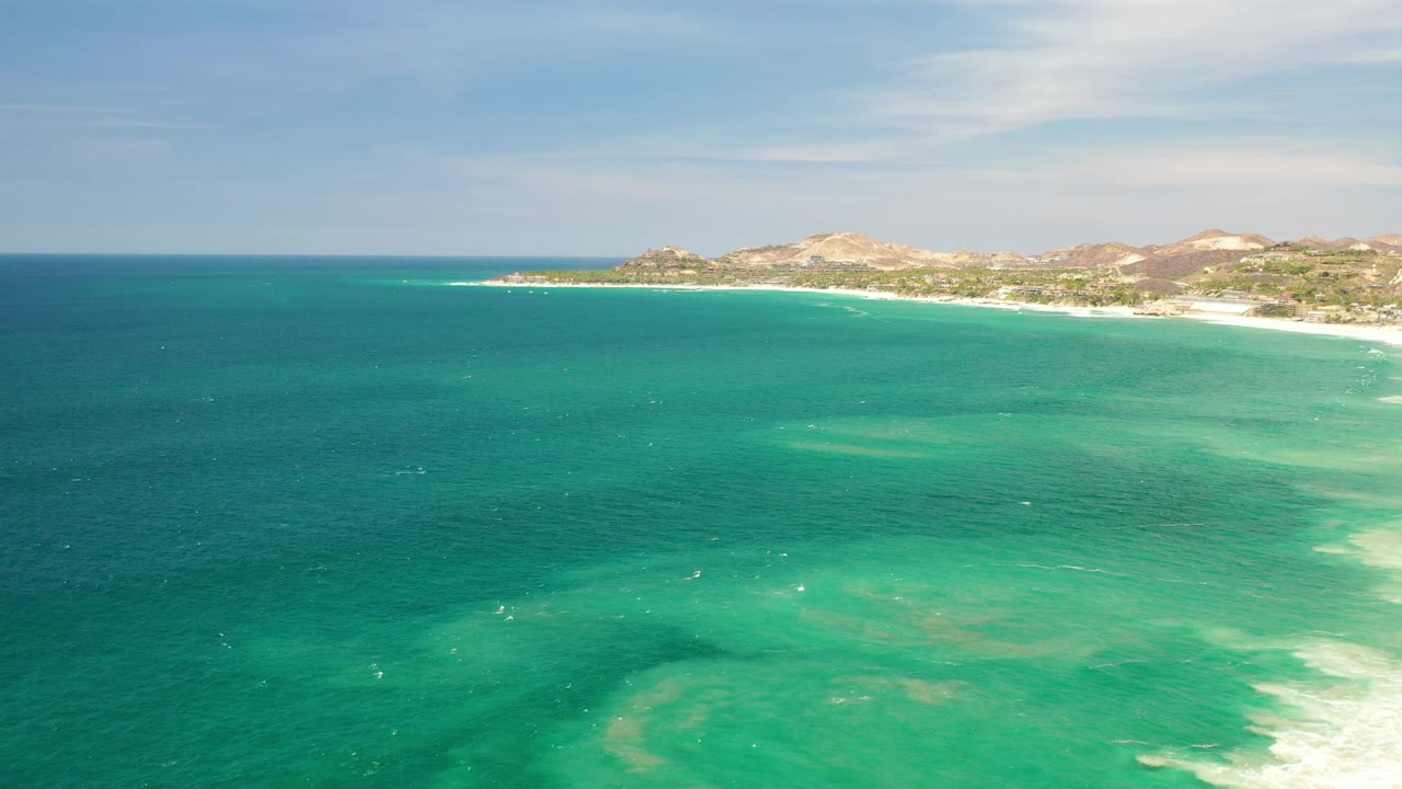 Opening aerial frame over Los Cabos, Mexico, with turquoise choppy water in front, a pale surf line along the right shore, low tan mountains on the horizon, and no visible people.