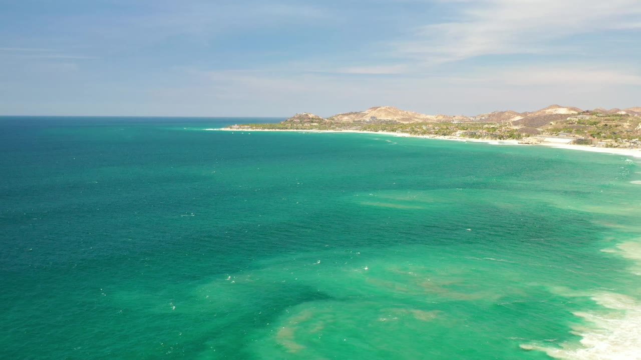 Aerial view in Los Cabos, Mexico, with dark teal water across the left foreground, lighter turquoise shallows near the right surf, distant dry hills, and no visible people.