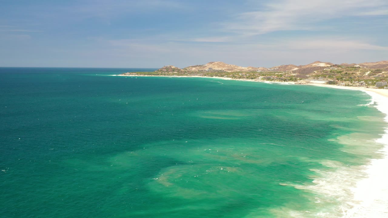 Opening aerial view over Los Cabos, Mexico coastline with teal ocean in the foreground, white surf on the right, low hills on the horizon, and no visible people.