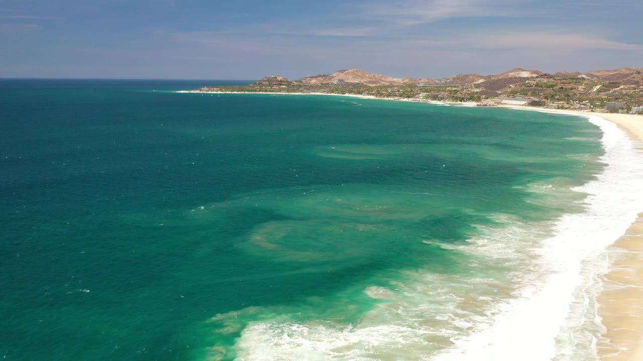 Aerial view of blue ocean, surf, beach, and distant low mountains.