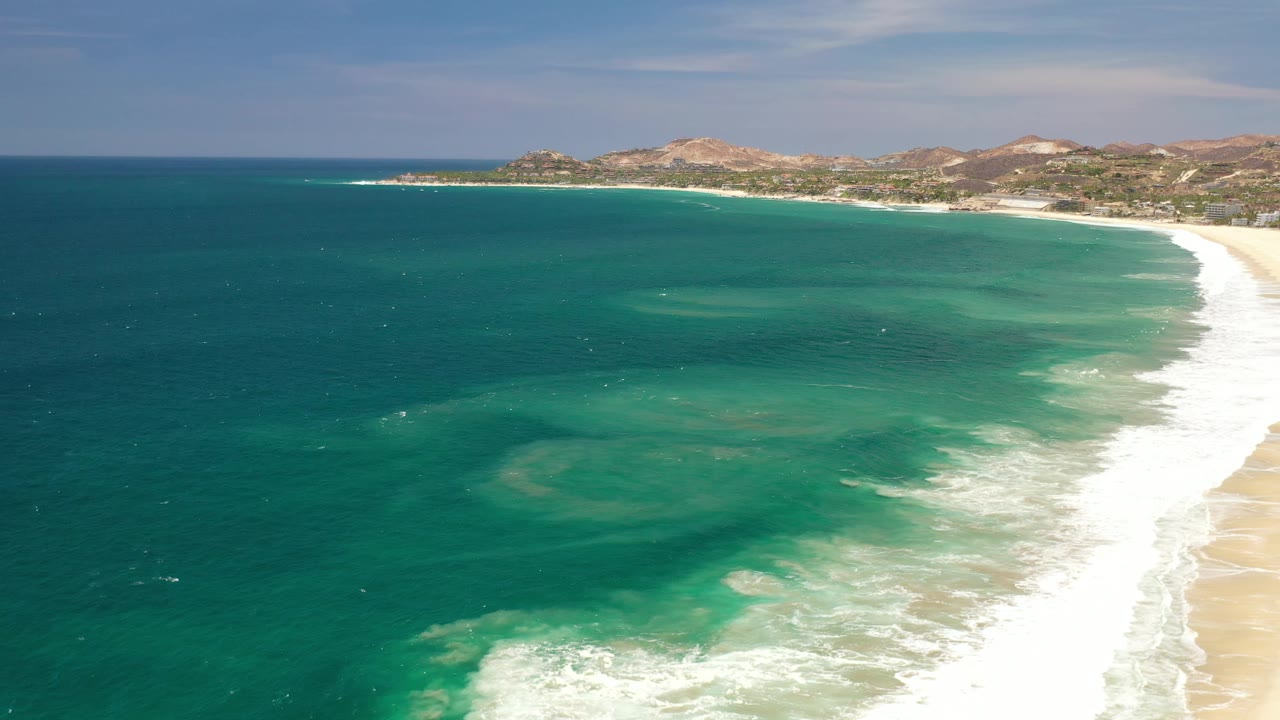 The Los Cabos shoreline remains wide in frame, with darker offshore water, lighter shallows near the surf, sand on the right, hills on the horizon, and no visible people.