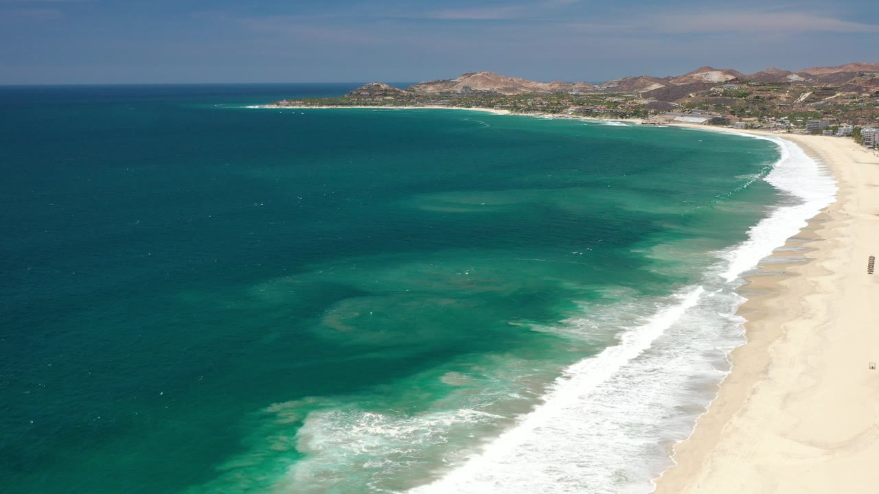 Opening aerial frame over Los Cabos, Mexico, with teal ocean on the left, a pale beach on the right, white surf along the shoreline, distant hills, and no visible people.
