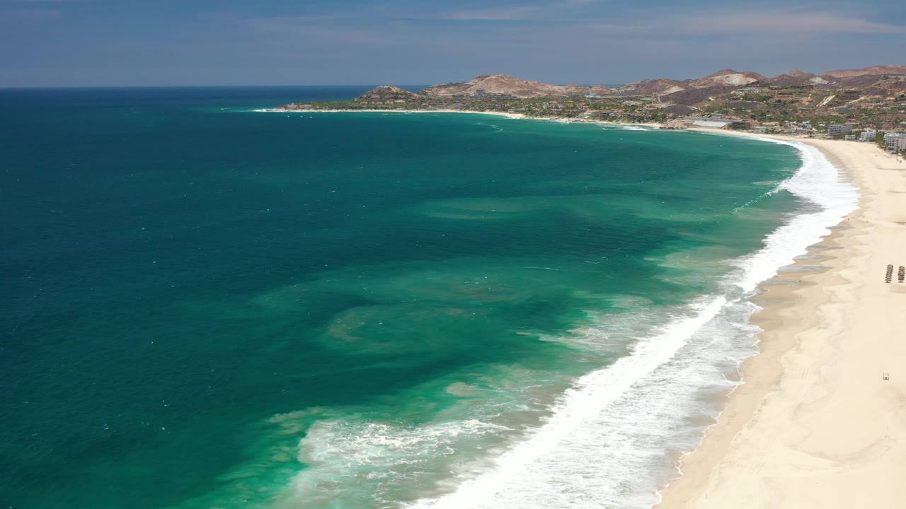 Aerial beach view with turquoise shallows, foamy surf, sand, and low hills.