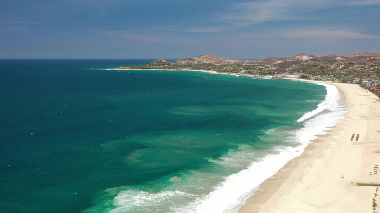 Opening frame of Los Cabos, Mexico from above, with deep blue ocean at left, a white surf line curving along pale sand, distant dry hills, and no visible people.