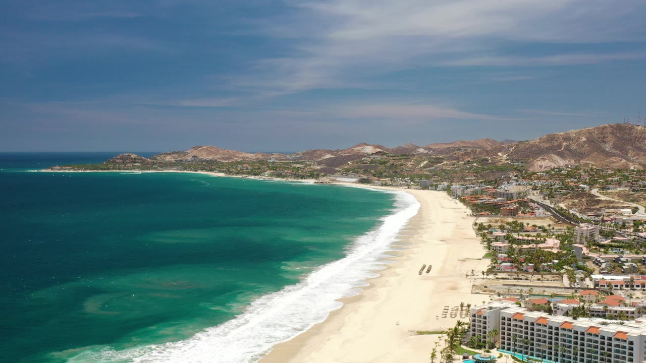 Wide aerial view of ocean, surf, sandy beach, hotels, and hills.