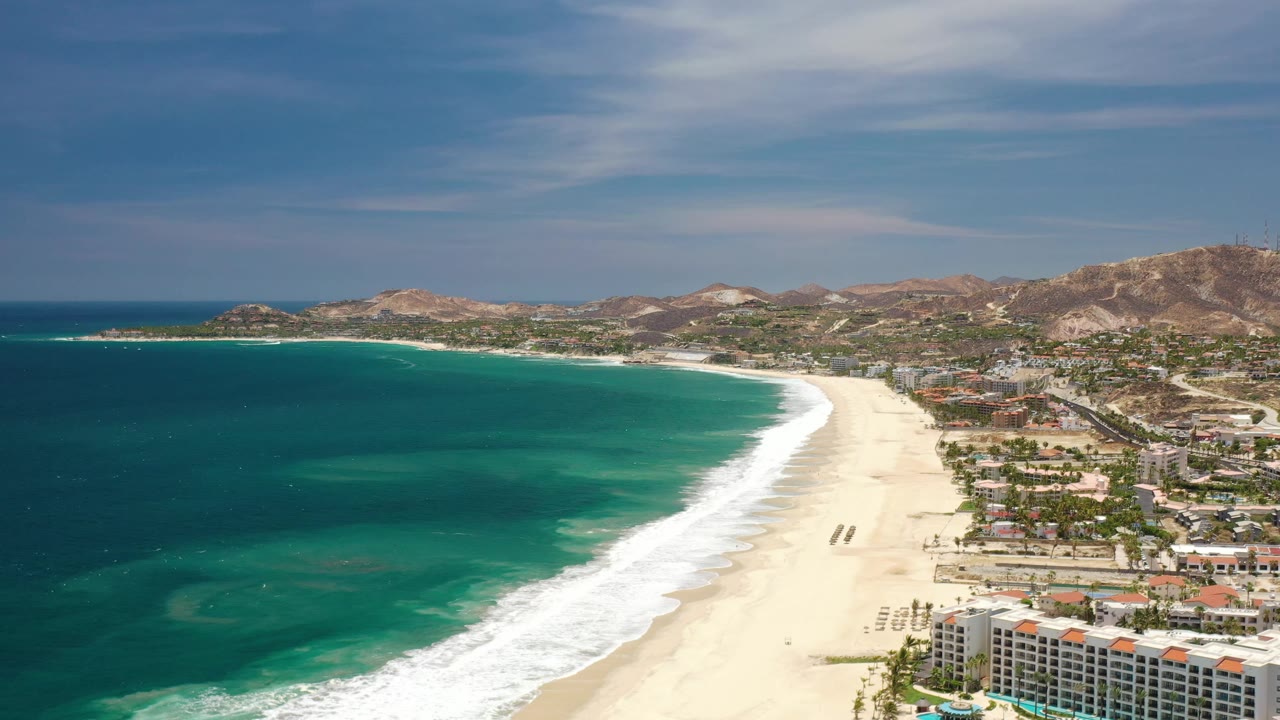 Los Cabos aerial frame showing a curved white surf line, pale sand, teal water, resort buildings near the lower edge, and hillside neighborhoods beyond the beach.