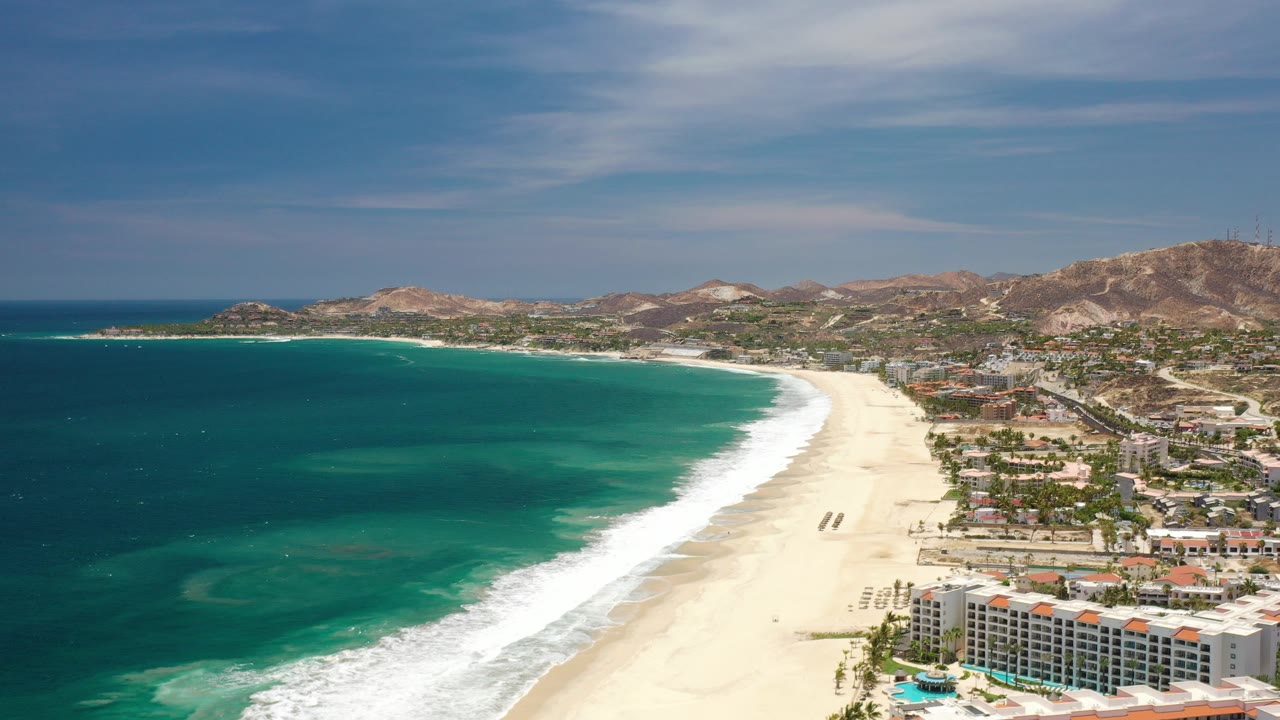 Opening aerial frame over a Los Cabos, Mexico beach, with turquoise water on the left, a bright sand shoreline, resort buildings in the lower right, hills beyond, and no visible people.
