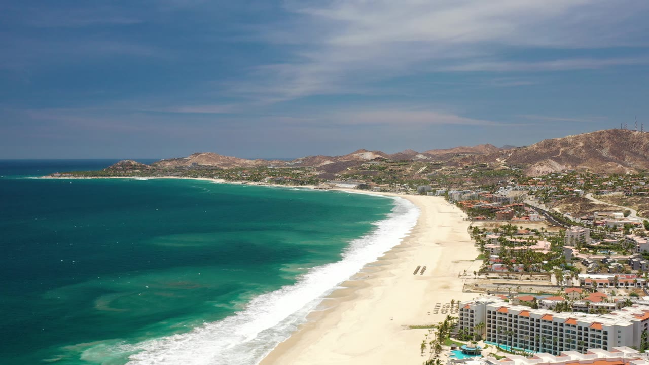 Aerial beach resort coastline with bright surf, ocean shallows, and hills in back.