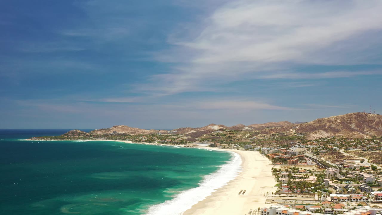 Aerial view in Los Cabos, Mexico, showing a broad band of turquoise shoreline, white surf on pale sand, the coastal town to the right, brown hills in back, and small distant figures on the beach.