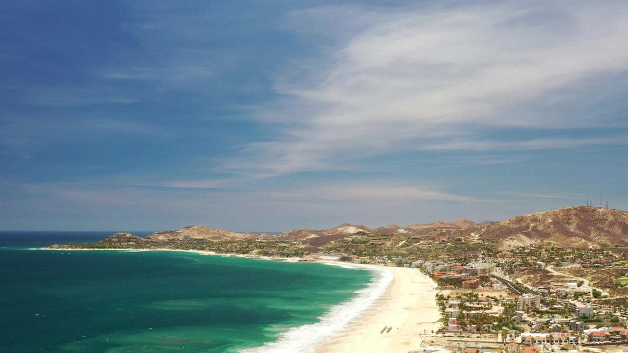 Aerial view of turquoise ocean, sandy beach, resort buildings, and dry hills.