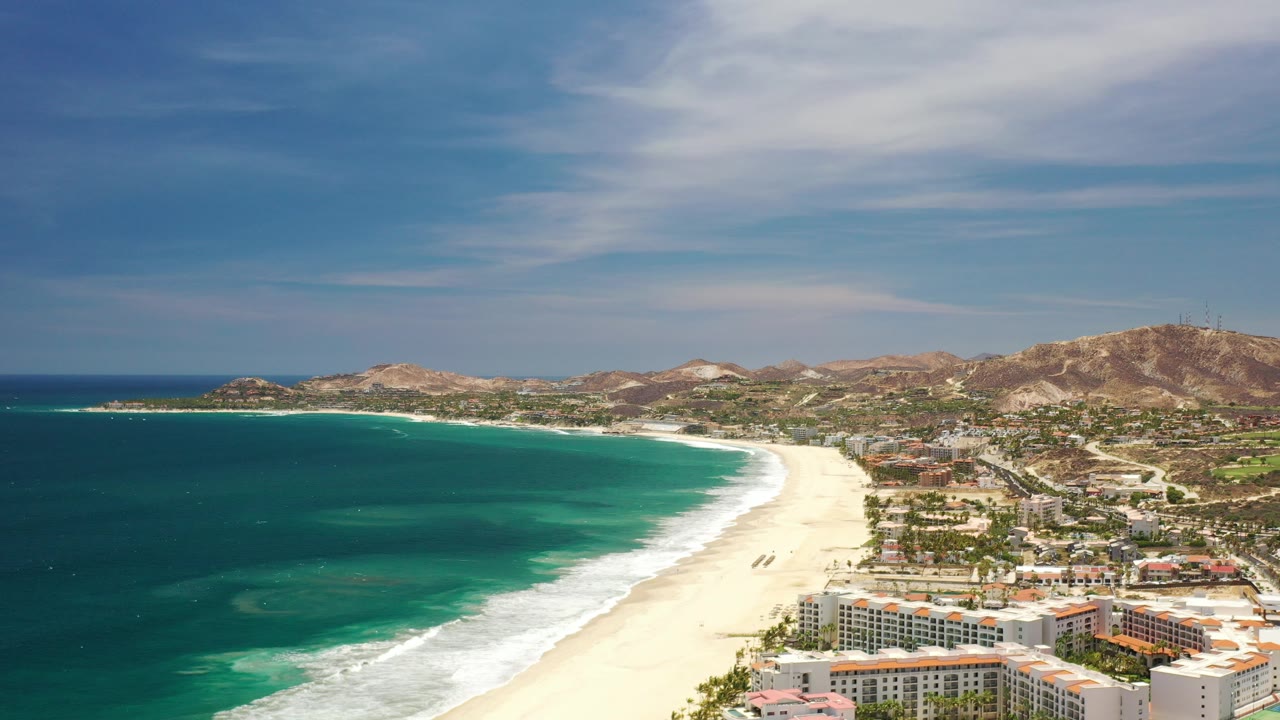 Wide aerial frame of the Los Cabos coastline with turquoise water on the left, a pale sandy beach curving through the center, coastal buildings below, dry hills beyond, and no visible people.