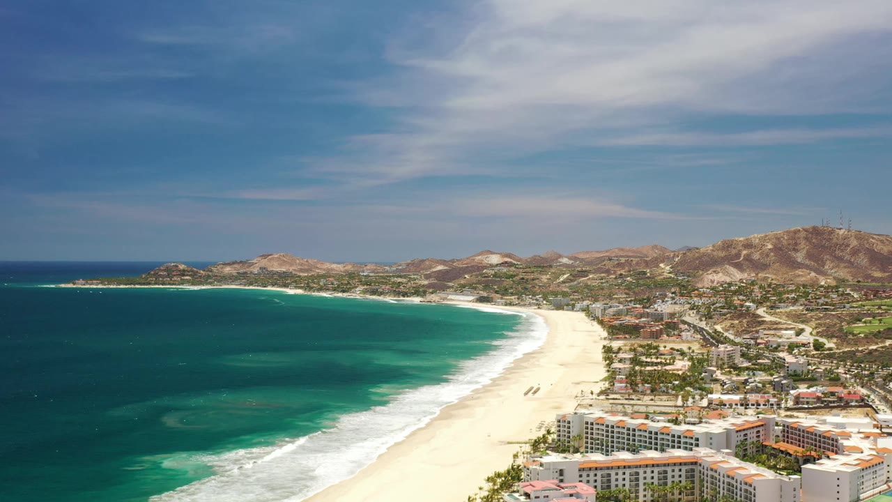 Los Cabos aerial view with the surf line bright against the sand, clustered coastal buildings in the lower frame, roads leading inland, brown hills across the background, and no visible people.