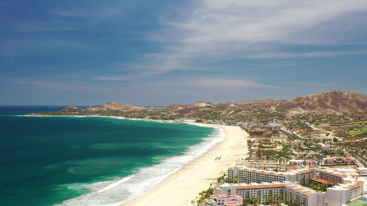 Turquoise bay, curving beach, resort-like buildings, and arid hills.