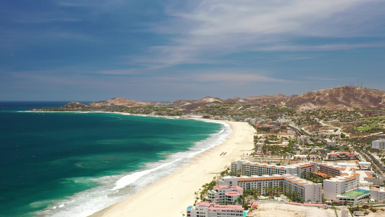 Opening aerial frame over Los Cabos, Mexico, showing a broad sandy beach beside dark blue and turquoise water, resort buildings in the foreground, inland hills beyond, and no visible people.