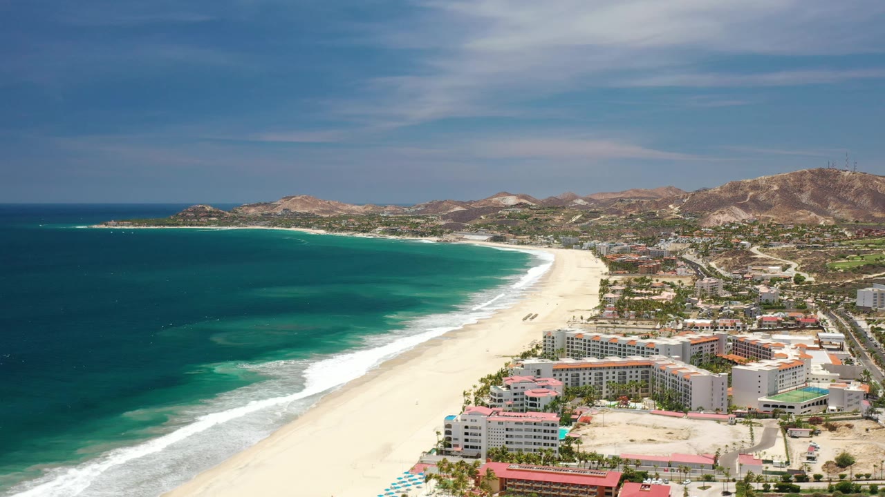 Los Cabos aerial frame showing the long white beach and turquoise water, with hotel blocks and roads in the lower right and dry hills across the background.