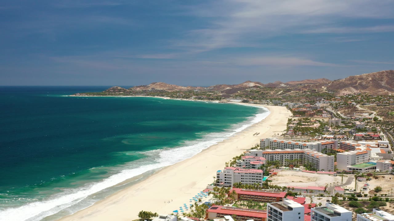 Opening aerial frame over Los Cabos, Mexico, with teal ocean at left, a pale beach curving past beachfront buildings, dry hills beyond, and no visible people.