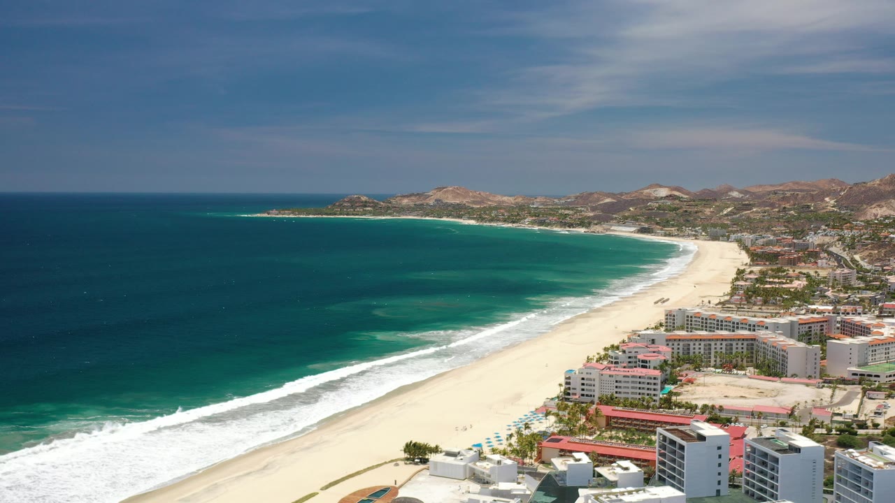 High aerial view over Los Cabos, Mexico with deep blue ocean on the left, a pale sand beach curving past shoreline buildings, dry hills beyond, and no visible people.