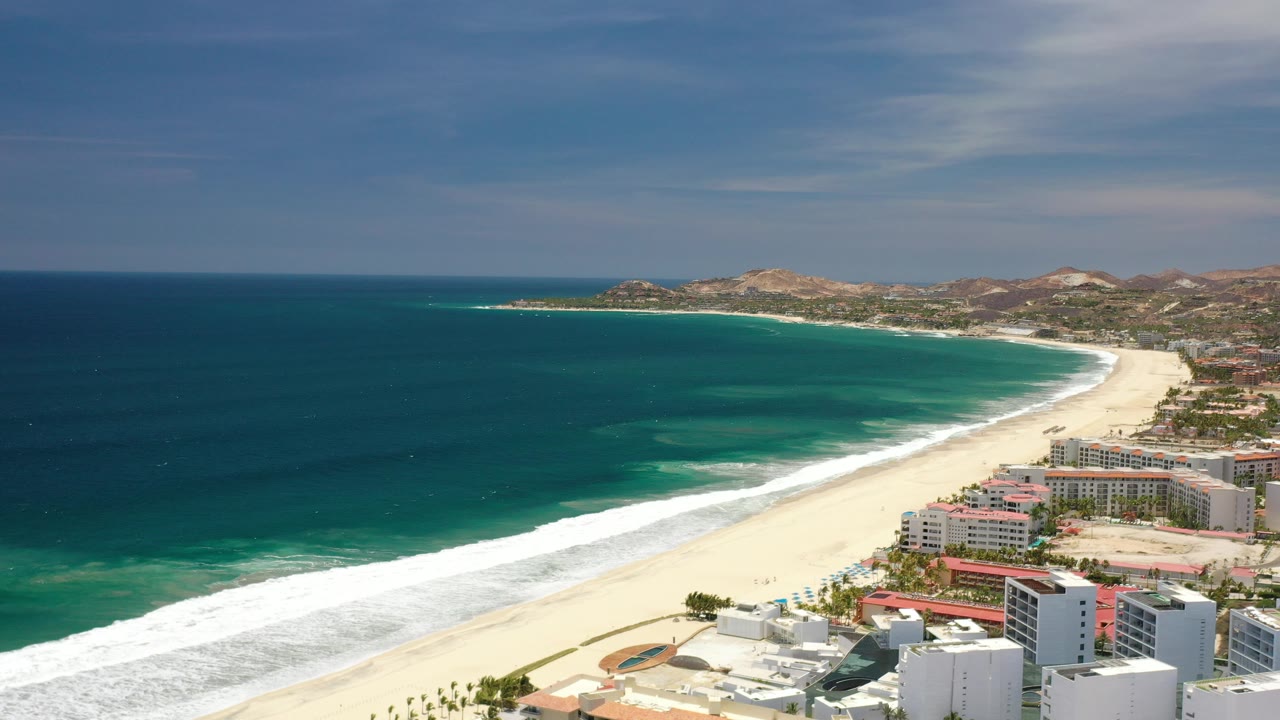 High aerial frame over Los Cabos, Mexico, showing deep blue and green ocean, a long white surf line, pale sand, beachfront hotel buildings, low hills, and no visible people.