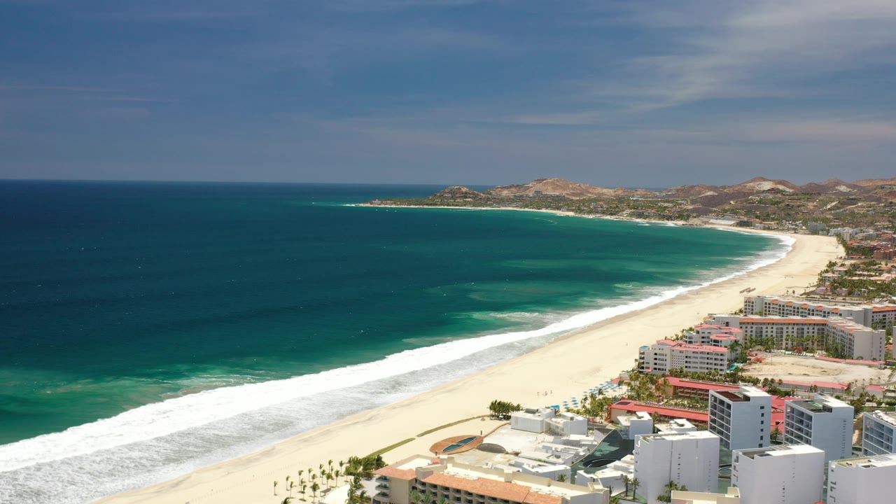 Aerial beach scene with curved shoreline, green water, and hotels at frame edge.