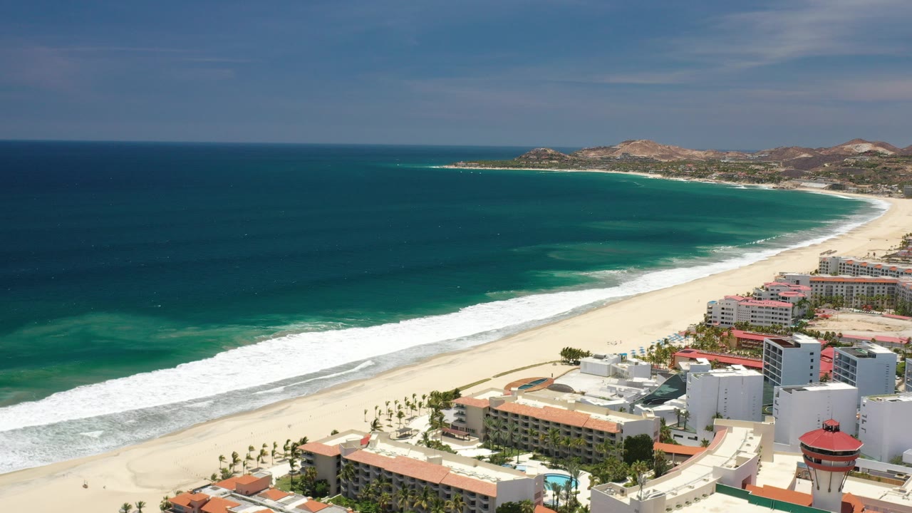 Aerial view over Los Cabos, Mexico, with dark blue ocean, a white surf line, sandy beach, and beachfront buildings in bright sun, with no visible people.