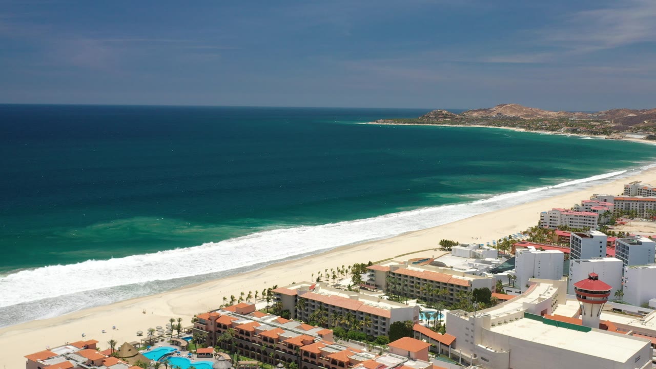 Aerial view over Los Cabos, Mexico showing dark blue ocean, a white surf band, pale beach, beachfront hotels, pools, palms, and dry hills in the distance with no recognizable people.