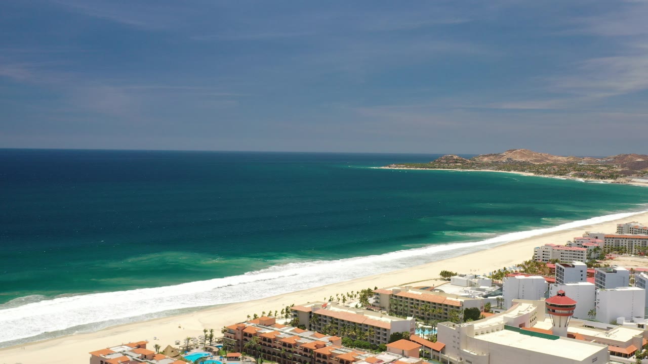 High aerial view over Los Cabos, Mexico resort hotels, courtyards, pools, a broad sand beach, turquoise ocean, and distant hills, with no visible people.