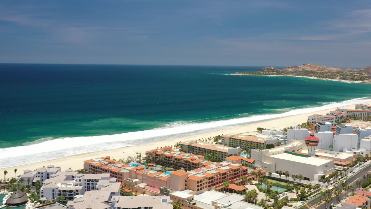 Opening aerial frame of Los Cabos, Mexico with deep blue ocean, a white surf line, coastal hotels in the foreground, distant dry hills, and no visible people.