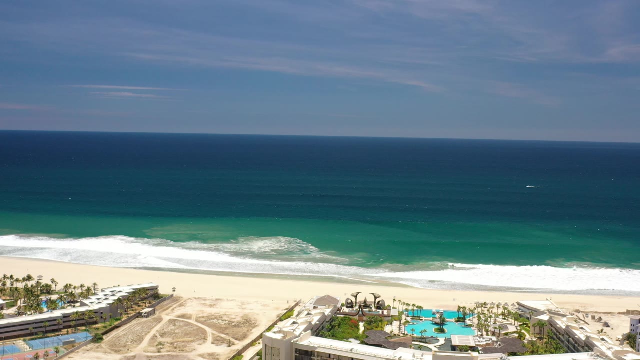 Opening Los Cabos, Mexico aerial frame with deep blue ocean, a turquoise nearshore band, white surf, pale sand, resort pools at the lower edge, and no visible people.