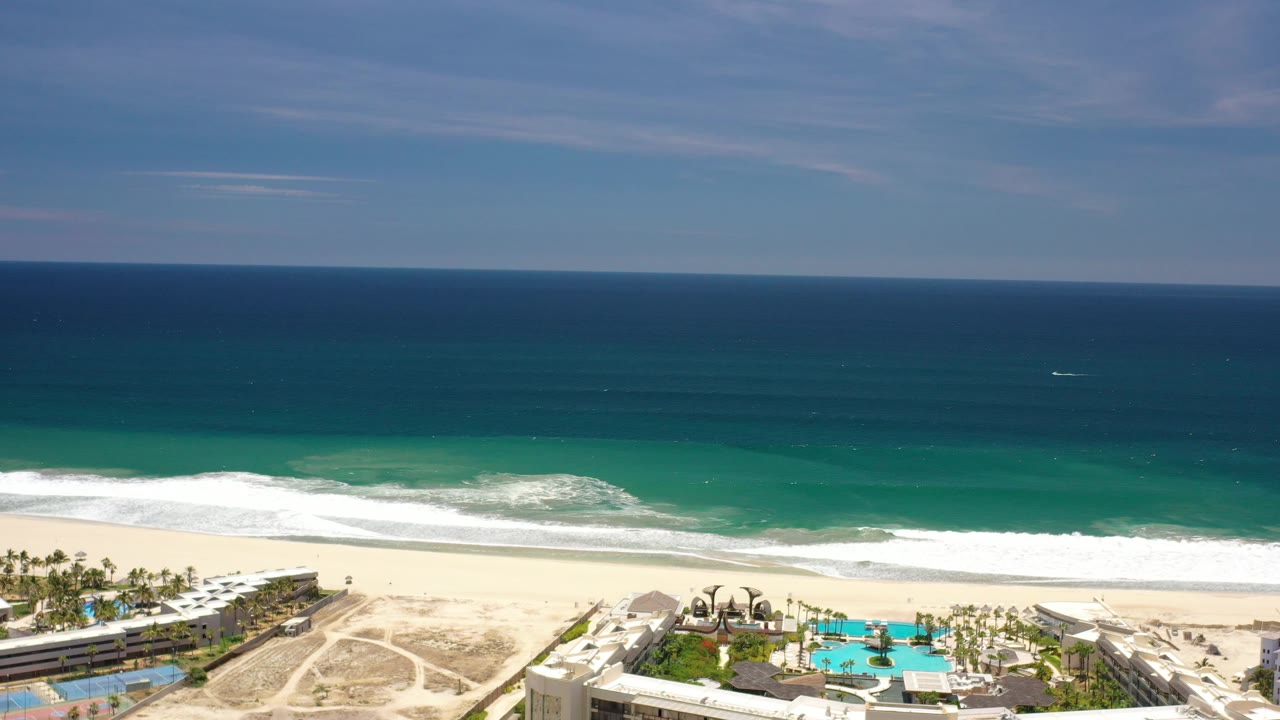High view over resort pools, sandy beach, white waves, and open sea.