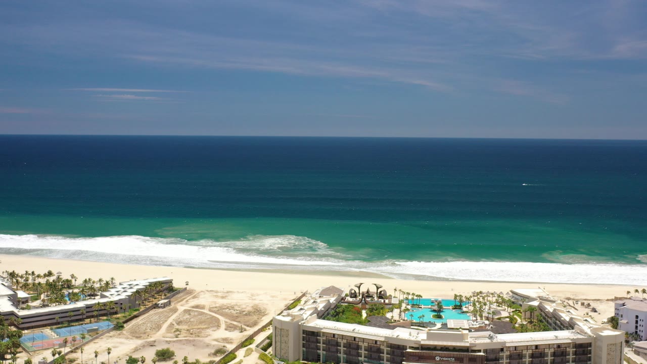 Opening frame of Los Cabos, Mexico from a high vantage point, with resort buildings and a turquoise pool at the bottom, pale sand and surf beyond, deep blue water, light clouds, and no visible people.